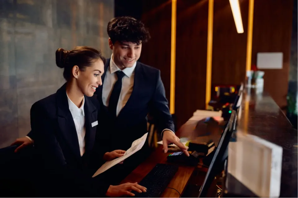 Two receptionists at a hotel front desk, one pointing at the screen